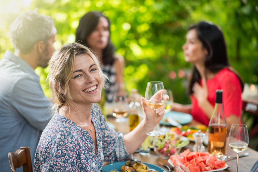 Looking at camera, portrait of a beautiful middle aged blond woman sharing a meal friends on a terrace table in summer, she has a glass of wine at hand. Shot with flare