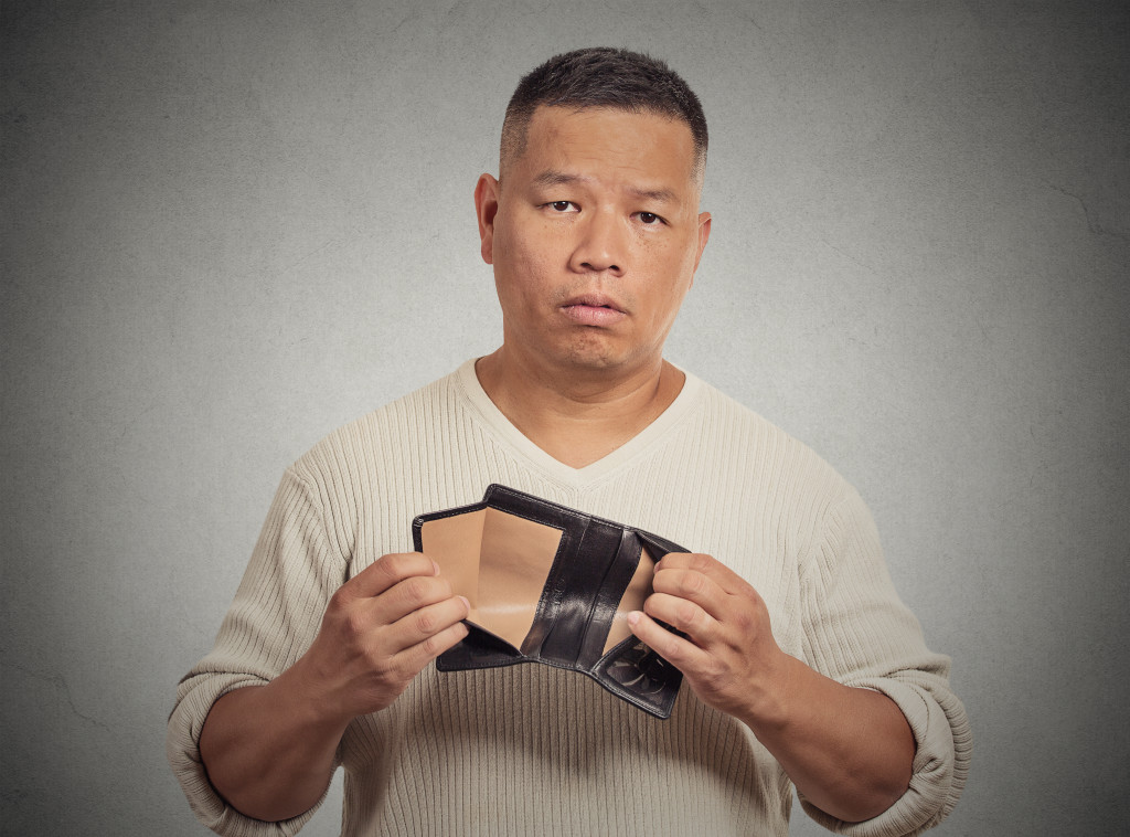 Closeup portrait stressed, upset, sad, unhappy middle aged man standing with, holding empty wallet isolated grey wall background
