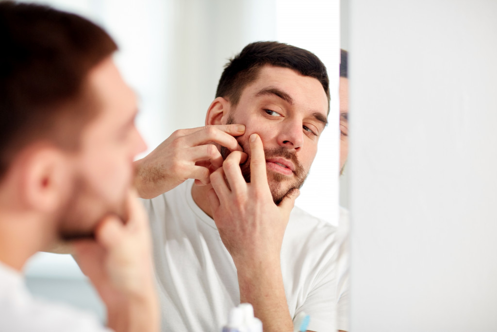 A man squeezing a pimple on his cheek in front of a mirror