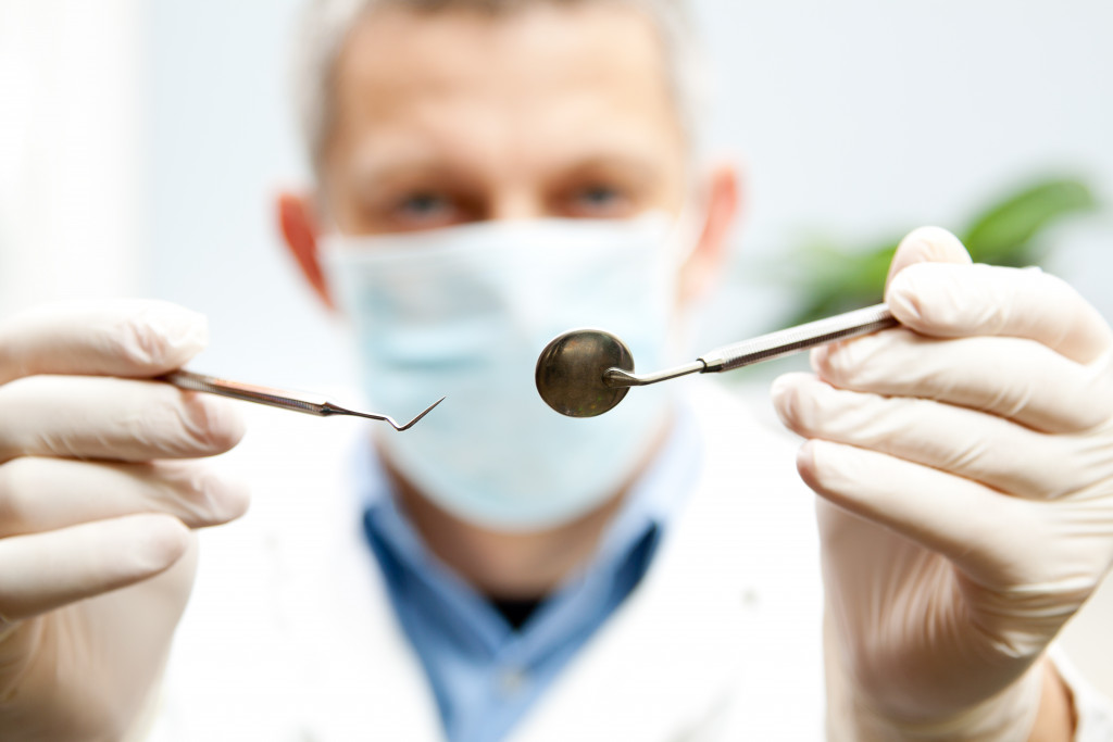 Focus on dentist's hands holding dental mirror and hook in the foreground, with the dentist in the background wearing a mask