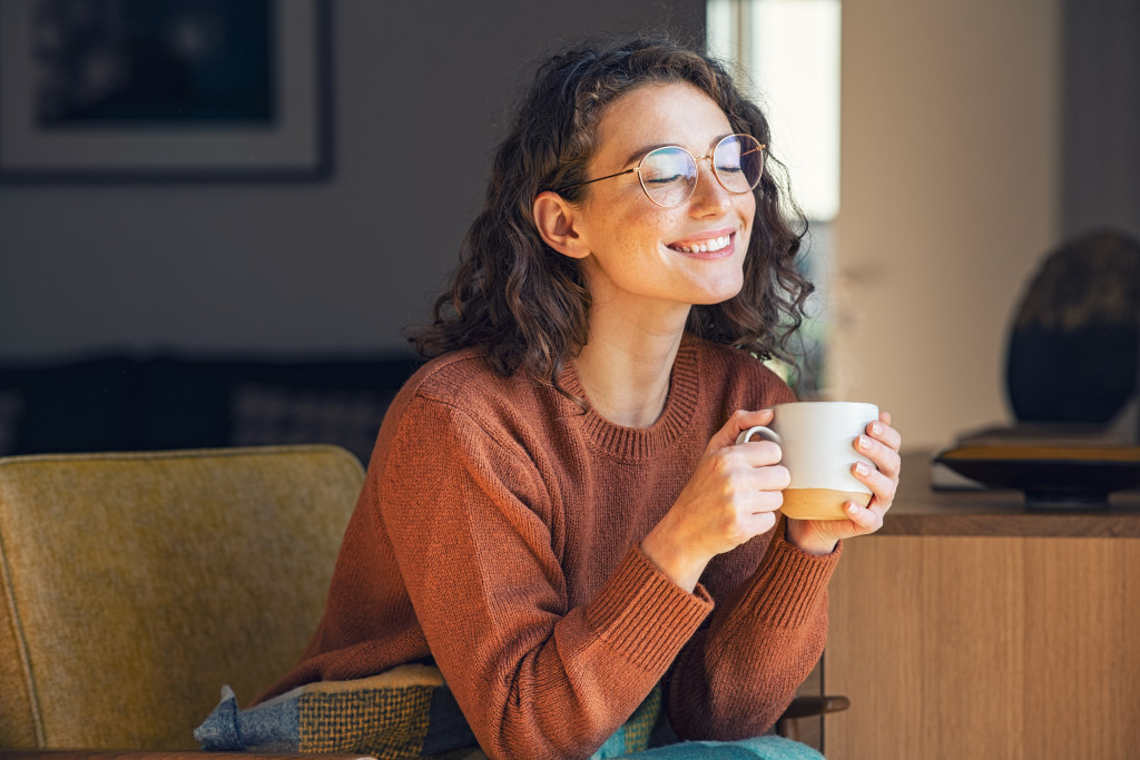 Smiling young woman enjoying the warm temperature inside a house while holding a cup of tea during winter.